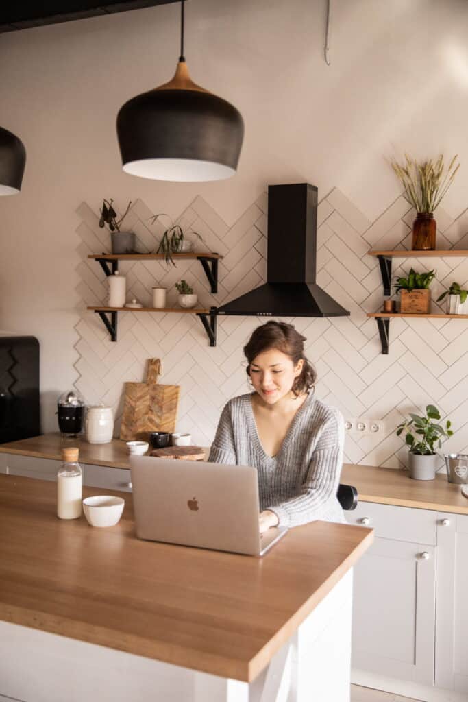 Woman working on laptop in modern kitchen with shelves and plants.